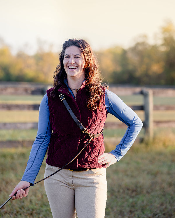 A woman laughs. She is wearing a corduroy vest, and across her shoulder is slung a leather dog harness. In her right hand is a leather dog leash.