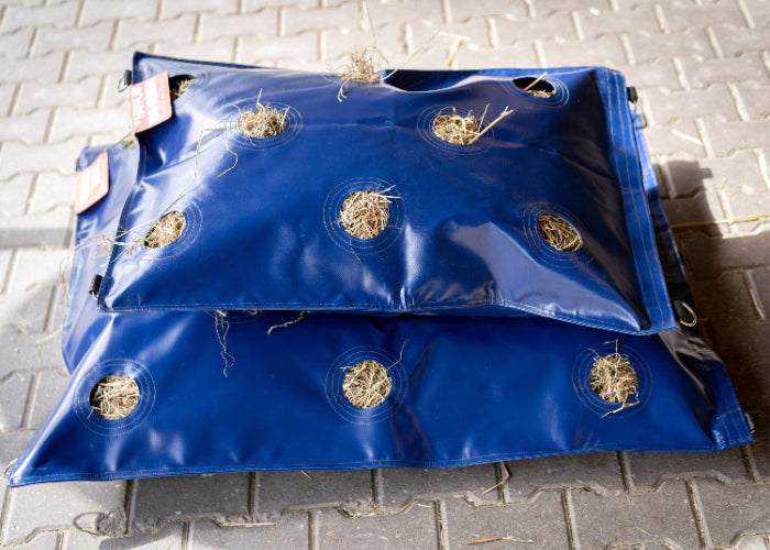Medium hay bag sitting on top of XL hay bag filled with hay on a barn floor
